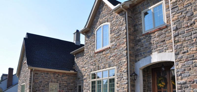 Two-story house with exterior walls made of decorative stone, large windows with white frames, dark shingled roof, and a front door decorated with a wreath. The sky is clear and blue.