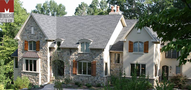 Two-story house with stone and stucco exterior, arched windows, wooden shutters, and a steep gray roof, surrounded by trees; Monogram Custom Homes logo in the top left corner.