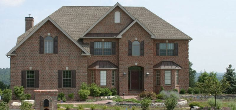 Two-story brick house with brown shutters, a gabled roof, several large windows, and landscaped front yard with shrubs and trees.