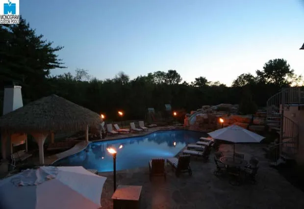 A backyard pool area at dusk features lounge chairs, umbrellas, tiki torches, a thatched-roof cabana, and trees in the background under a darkening sky.