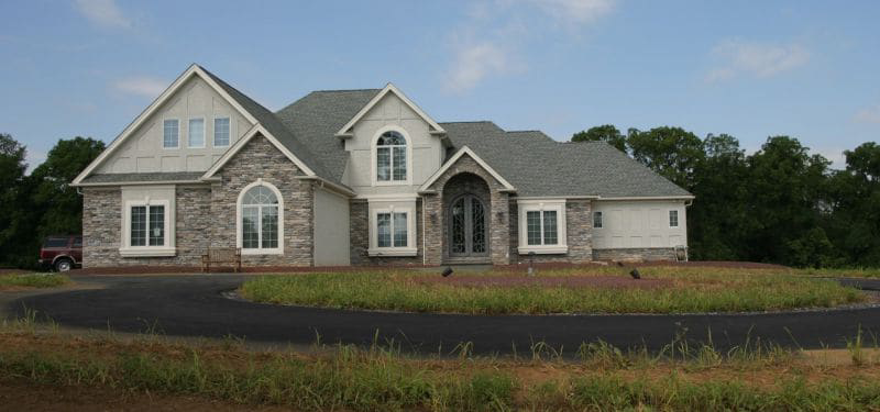 A large, two-story house by Monogram Custom Builders with gray stone and siding exterior, arched windows, and a three-car garage sits on a spacious lot with a circular driveway and grass, surrounded by trees under a partly cloudy sky.