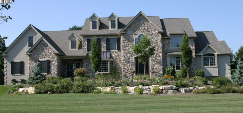Large two-story house with stone and beige siding, multiple gables, and dormer windows. Built by custom home builders, it features a well-maintained lawn, landscaped garden beds, shrubs, and small trees in the front yard.
