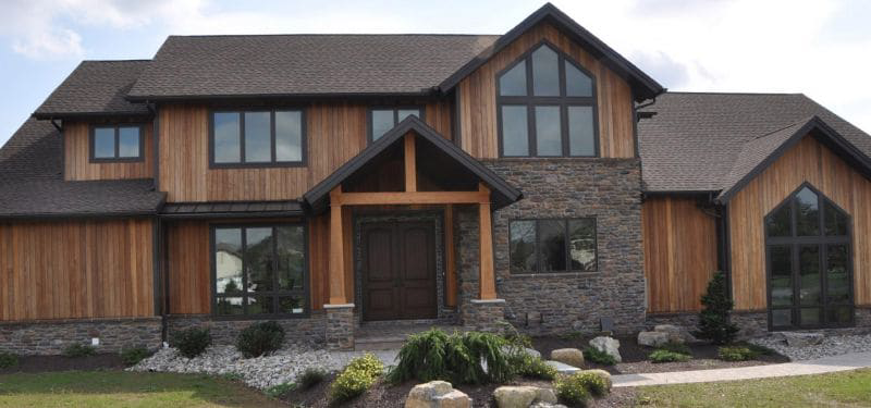 A modern two-story house with wood siding, stone accents, large windows, and a landscaped front yard featuring rocks and shrubs under a partly cloudy sky—perfect for those seeking stylish design with thoughtful specifications.