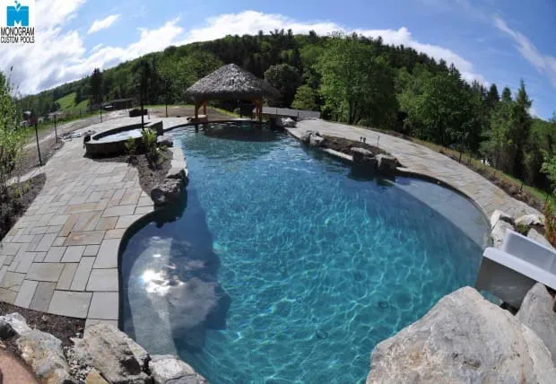 A landscaped backyard pool with clear blue water, surrounded by stone decking, natural rocks, a hot tub, and a thatched-roof gazebo, set against a backdrop of trees and rolling hills.
