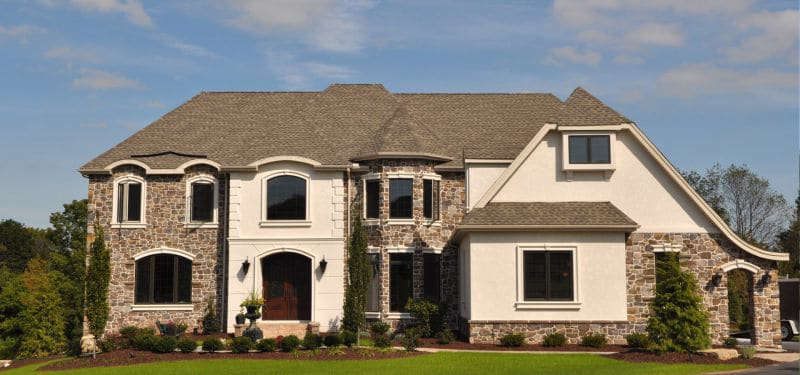Large two-story house with stone and stucco exterior, arched windows, and a manicured lawn with shrubs and trees in front, under a blue sky with scattered clouds.