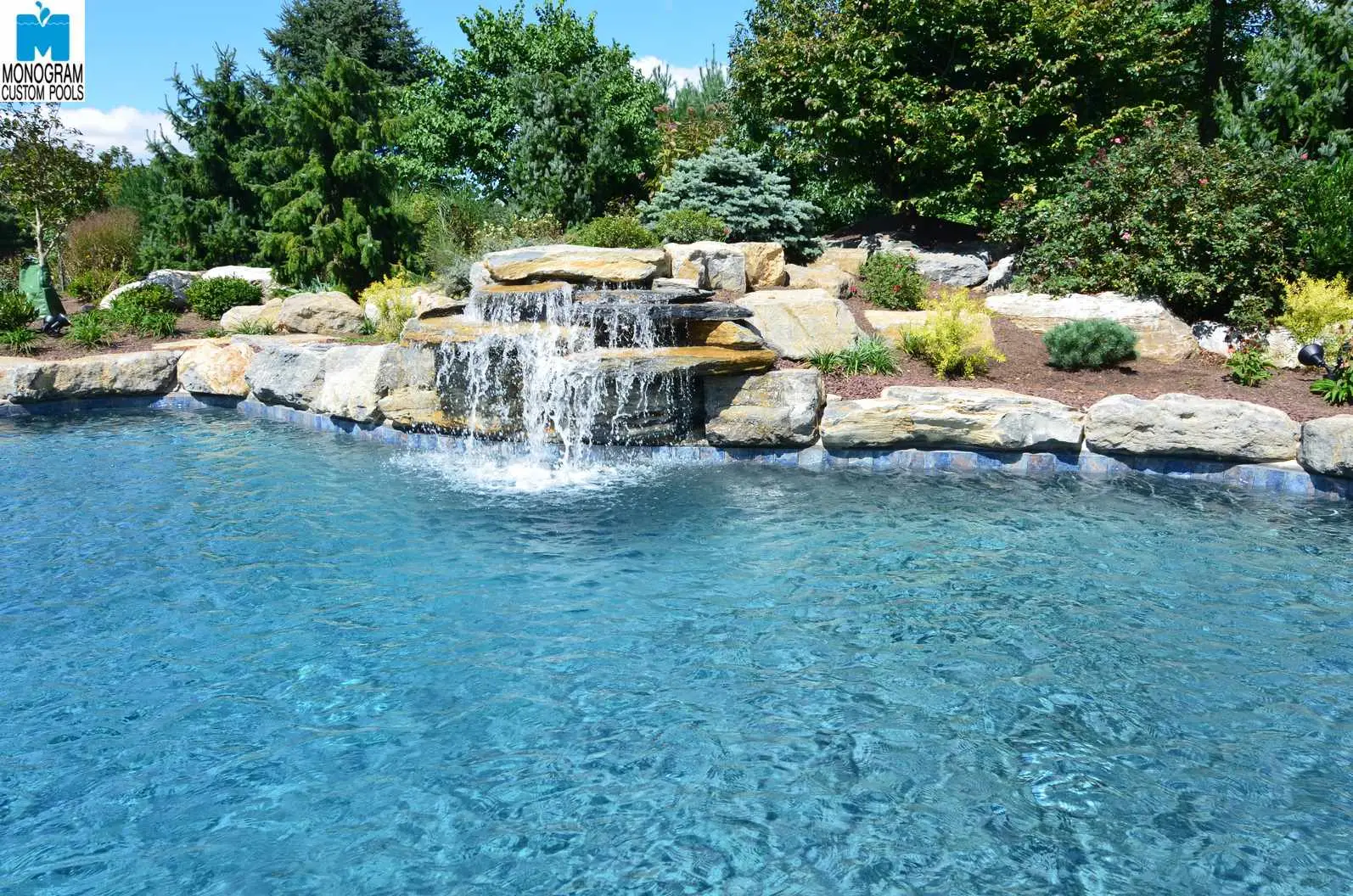 A clear blue swimming pool with a rock waterfall feature flows into the water, surrounded by landscaping with green trees, bushes, and large decorative rocks under a sunny sky.