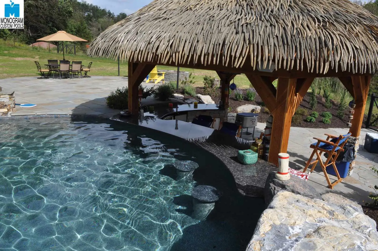 A backyard pool with clear water features a swim-up bar under a thatched-roof pavilion with wooden beams. There are stone stools in the water and patio seating in the background on a stone deck.