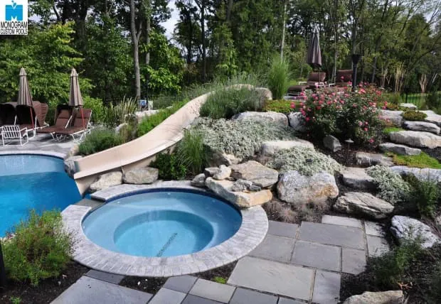 A landscaped backyard features a round hot tub next to a pool with a curved slide, surrounded by large rocks, flowering plants, patio chairs, and trees in the background.