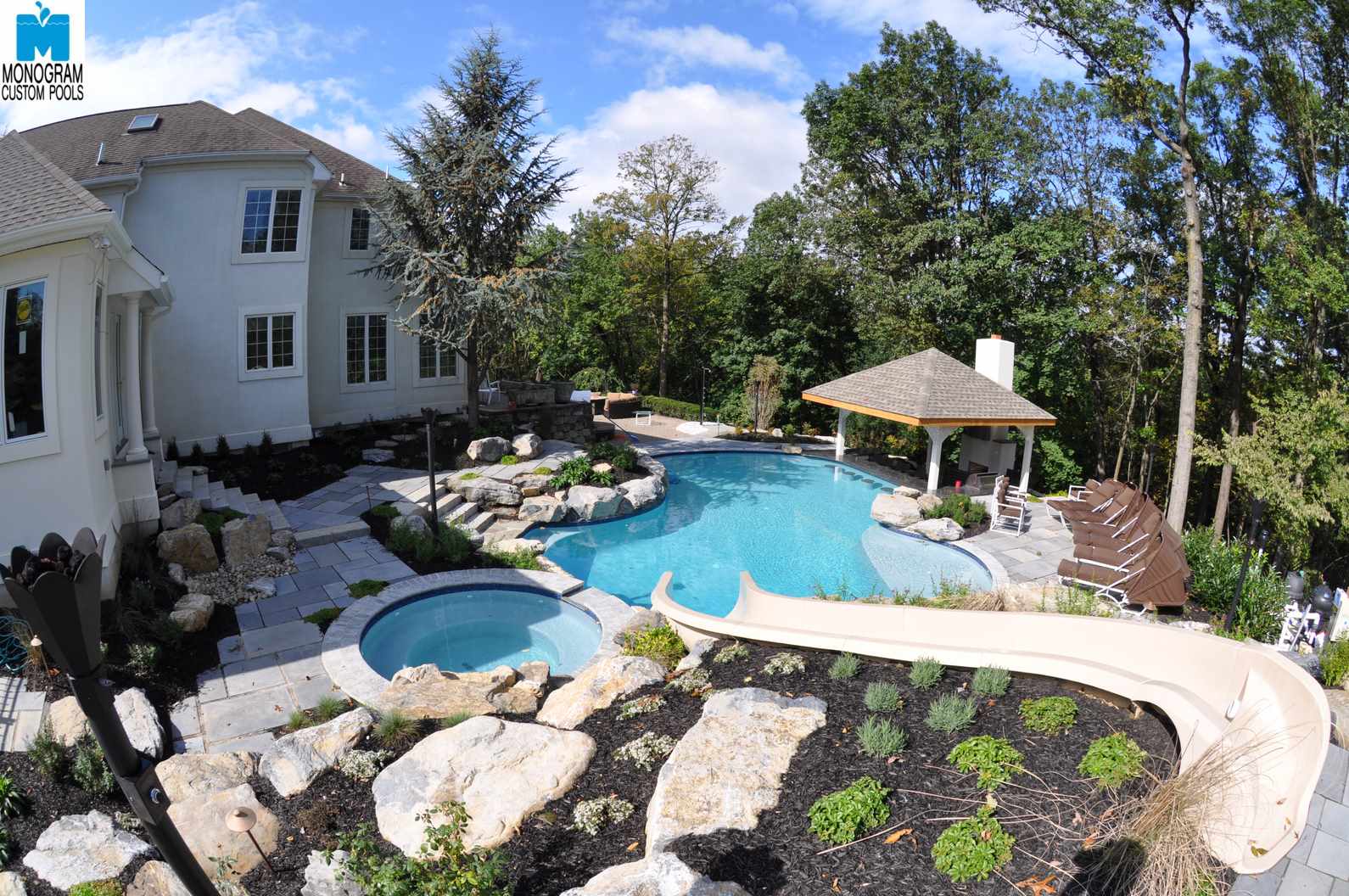 A luxurious backyard features a large, curvy swimming pool with a hot tub, rock landscaping, a water slide, patio seating, and a wooden pergola, surrounded by trees and a two-story white house.