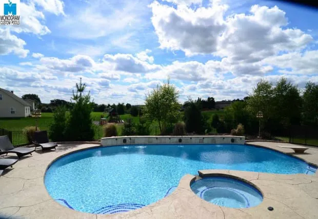 A backyard swimming pool with clear blue water, a connected circular hot tub, and surrounding patio. The pool overlooks a green lawn and trees under a partly cloudy blue sky. Lounge chairs are on the patio.