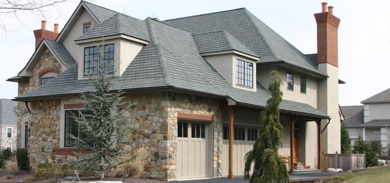 A two-story house with stone and stucco exterior, multiple peaked roofs, large windows, two brick chimneys, and a three-car garage—perfect to build On Your Lot. A few small trees and shrubs are in the front yard.