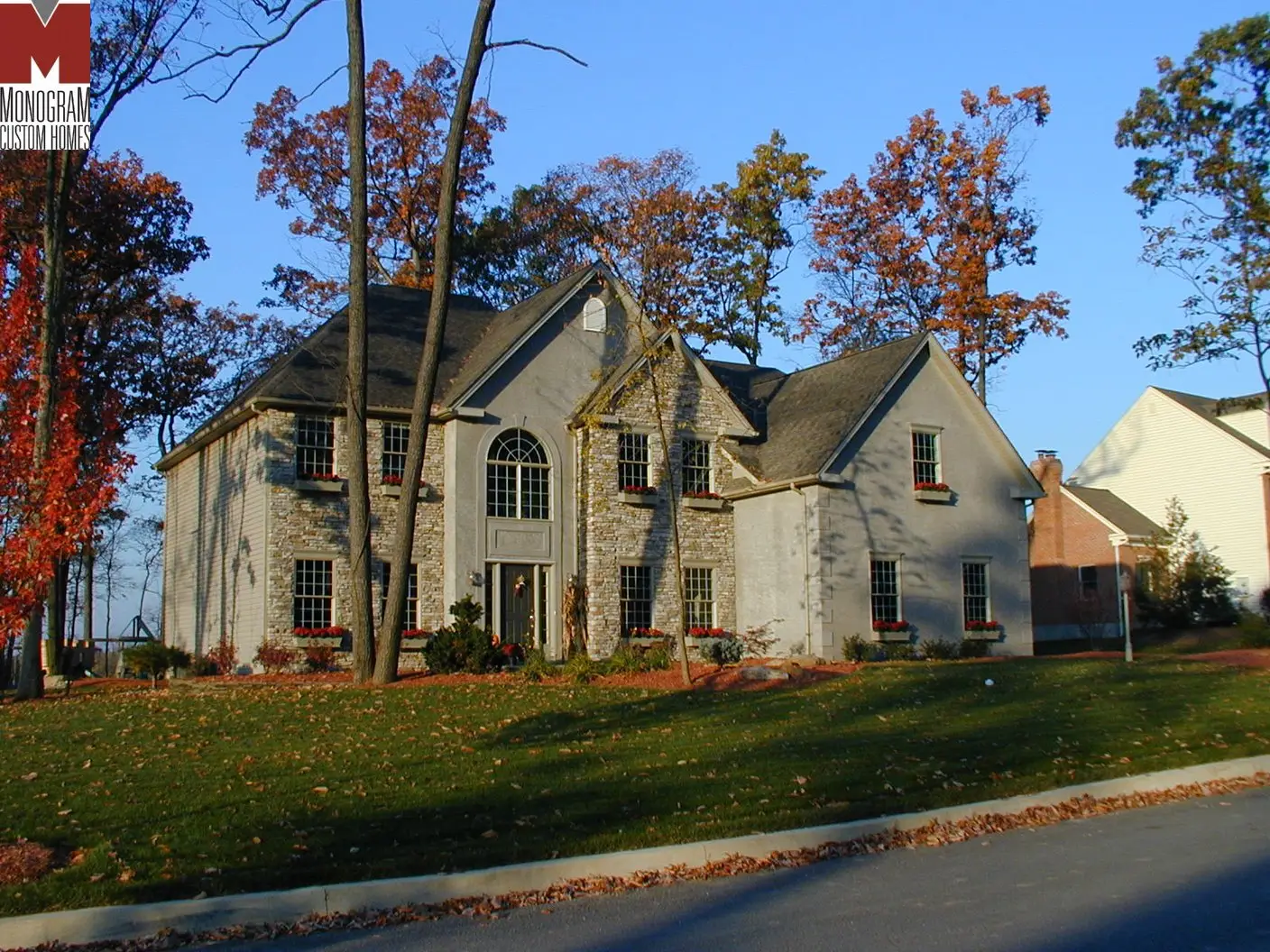 A two-story house with stone and light-colored siding, tall windows, and a sloped roof, surrounded by autumn trees and a grassy lawn, under a clear blue sky.