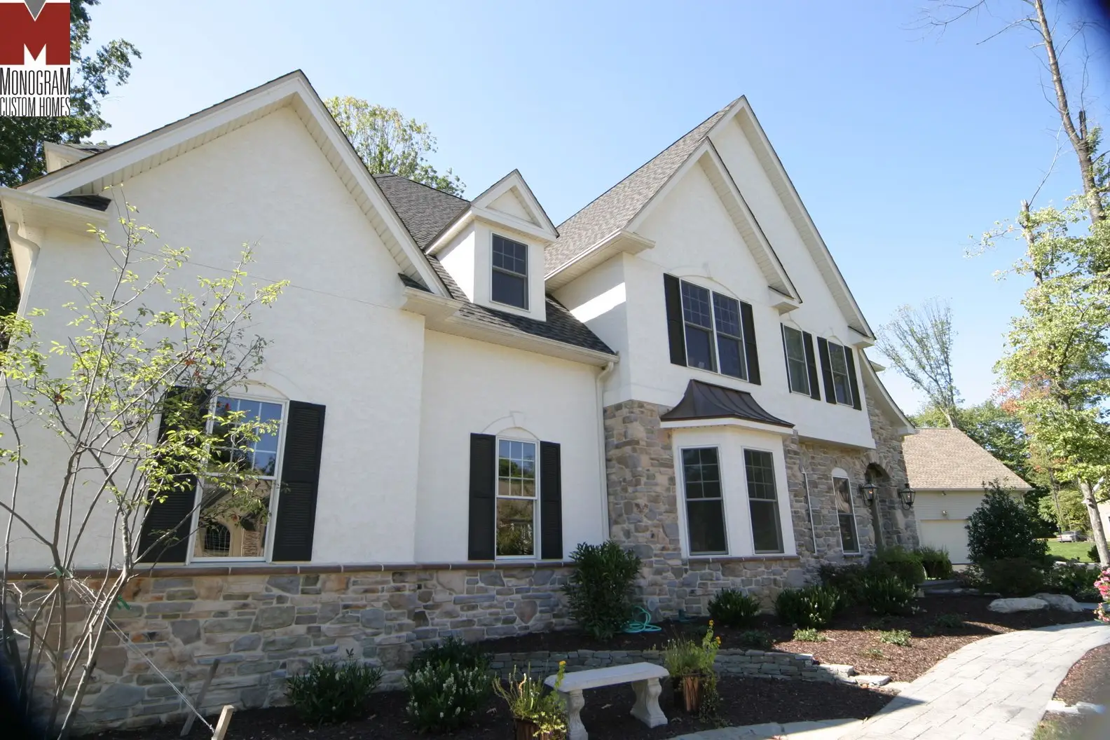 A two-story suburban house with cream stucco and stone exterior, dark shutters, large windows, gable roofs, and landscaped front yard with a small bench and pathway.