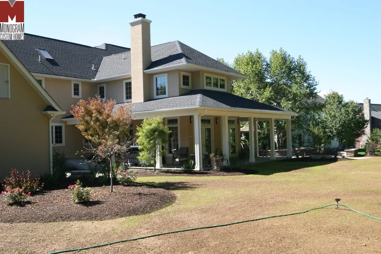 A large beige two-story house with a covered patio, many windows, and a landscaped yard with some trees and garden beds. A green garden hose lies on the grass in the foreground.