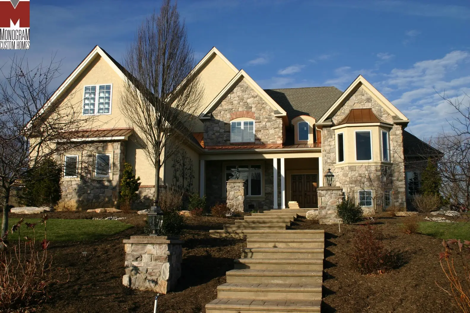 A large, two-story house with a stone and stucco exterior, steep gabled roofs, and multiple windows sits atop a landscaped yard with steps leading to the front entrance on a clear, sunny day.