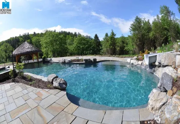 A backyard swimming pool with clear blue water, surrounded by stone tiles and landscaping, features a thatched-roof cabana and natural rock accents. Trees and greenery fill the background under a blue sky.
