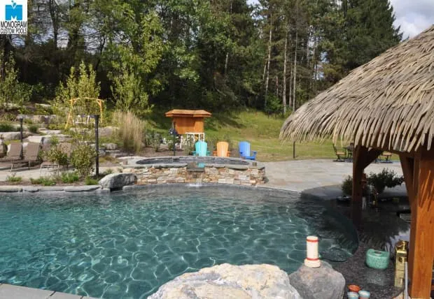 A backyard pool with rock accents, surrounded by trees and greenery. There’s a stone hot tub, colorful lounge chairs, and a tiki-style cabana with a thatched roof in the foreground.