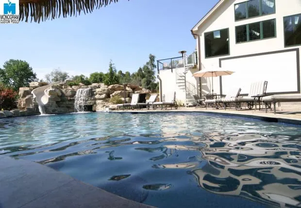 A backyard pool with clear water, surrounded by lounge chairs and umbrellas. A rock waterfall and water slide are visible in the background, next to a modern white house with large windows.