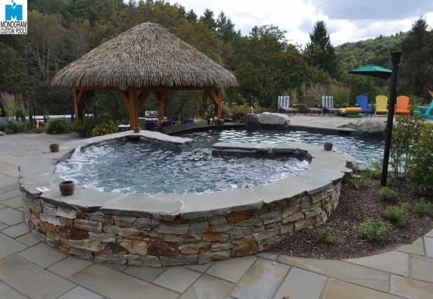 A round stone hot tub with bubbling water sits next to a lagoon-style pool. A thatched-roof gazebo and patio chairs with colorful cushions are in the background, surrounded by trees and greenery.