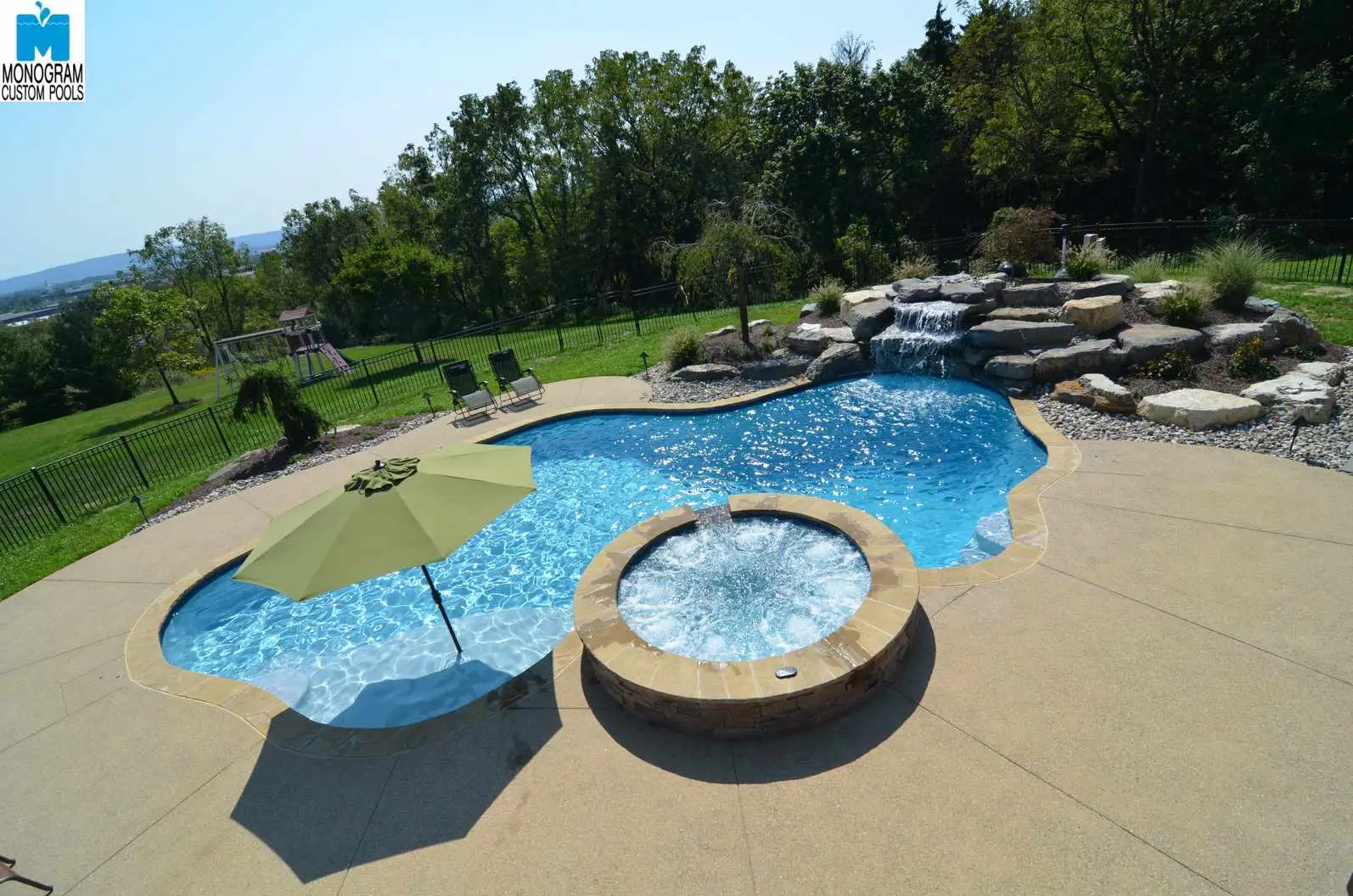 A backyard pool with an attached circular hot tub, a waterfall feature, lounge chairs, and a green umbrella, surrounded by a patio and grassy yard with trees and distant hills in the background.