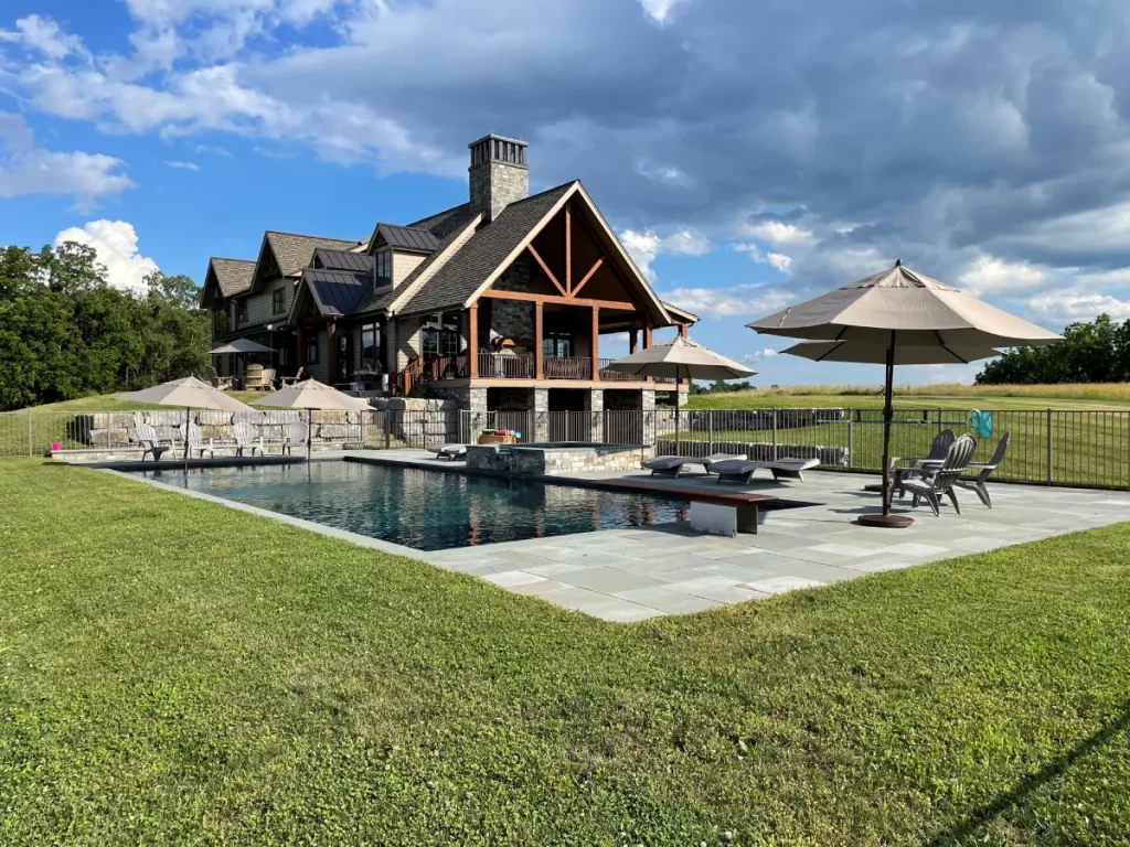 A large, rustic house with a stone chimney stands behind a fenced-in outdoor pool. Lounge chairs and umbrellas are arranged on the stone patio, surrounded by green grass and open fields under a partly cloudy sky.